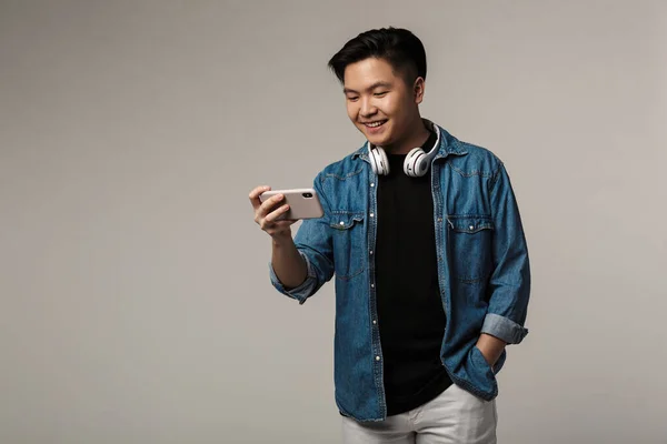 笑顔の若いアジア人男性のイメージは灰色の背景に隔離されたスマートフォンでビデオゲームをプレイ — ストック写真