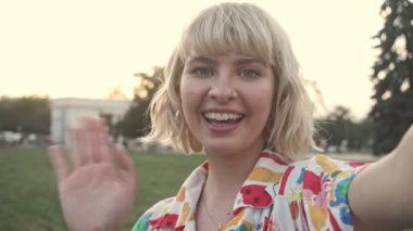 Çekici genç bir kadın, şehir parkında elini sallarken selfie videosu çekiyor.