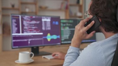 Ofiste oturmuş akıllı telefonuyla konuşan genç bir programcının arka planı bilgisayarla çalışıyor.