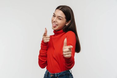 Beyaz duvarın üzerinde izole edilmiş, göz kırpan ve parmaklarını kameraya gösteren neşeli genç bir kadının fotoğrafı.