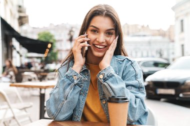 Gülümseyen güzel bir kadının görüntüsü. Cep telefonuyla konuşuyor ve açık hava kafede otururken kahve içiyor.