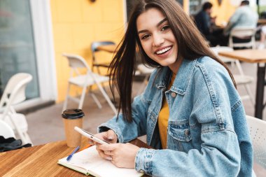 Gülümseyen güzel bir kadının sokak kafesinde otururken cep telefonu kullanması ve kahve içmesi.