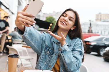 Gülümseyen bir kadının cep telefonuyla selfie çekmesi ve açık bir kafede otururken fotokopi çekmesi.