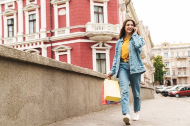 Kot pantolon giymiş genç bir kadının görüntüsü, şehir sokaklarında alışveriş çantalarıyla yürürken cep telefonuyla konuşması.