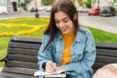 Şehir caddesinde bankta otururken gülümseyen ve not alan çekici mutlu bir kadın görüntüsü.