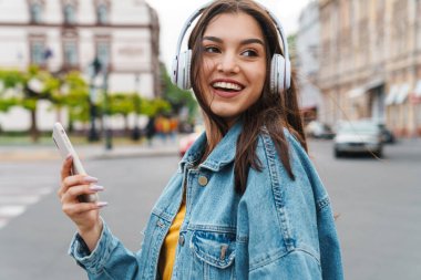 Akıllı telefon ve kablosuz kulaklıkla müzik dinleyen gülen güzel bir kadın görüntüsü.
