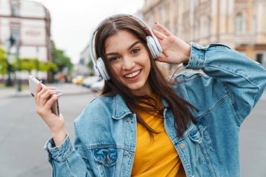 Akıllı telefon ve kablosuz kulaklıkla müzik dinleyen gülen güzel bir kadın görüntüsü.