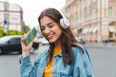 Akıllı telefon ve kablosuz kulaklıkla müzik dinleyen gülen güzel bir kadın görüntüsü.
