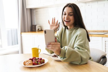 Modern mutfakta kahvaltı yaparken gülümseyen bir kadının el sallaması.