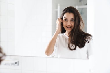 Banyoda aynaya bakarken gülümseyen esmer saçlı neşeli bir kadının fotoğrafı.