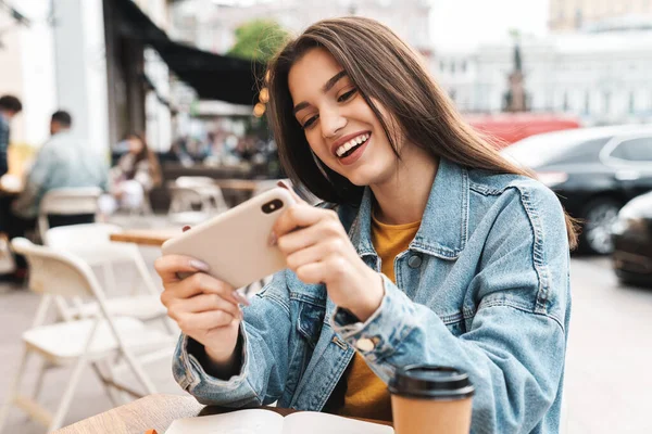 Image of joyful brunette woman playing video game on mobile phone while sitting at street cafe outdoors