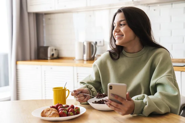 Fénykép Mosolygós Gyönyörű Használja Mobiltelefon Miközben Reggeli Modern Konyhában — Stock Fotó