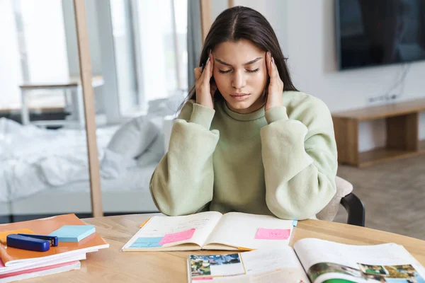 Photo of unhappy student woman with headache rubbing her temples while doing homework in living room