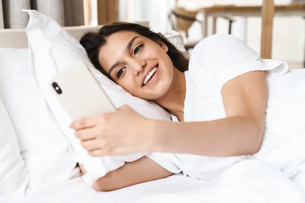 Фото Счастливой Женщины Улыбающейся Пользующейся Смартфоном Лежащей Постели Белой Подушке — стоковое фото
