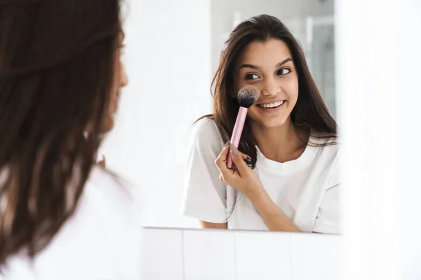 バスルームで鏡を見ながら笑顔とパウダーブラシを使用して幸せな素敵な女性の写真 — ストック写真