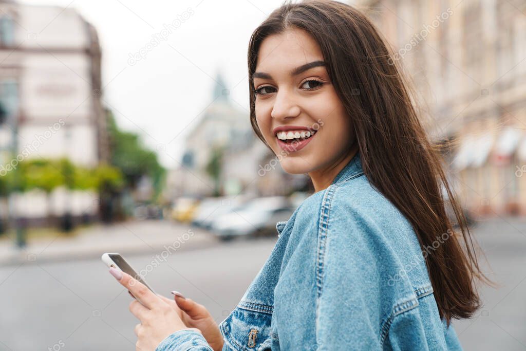 Image of attractive happy woman in denim jacket smiling and using smartphone while walking on city street