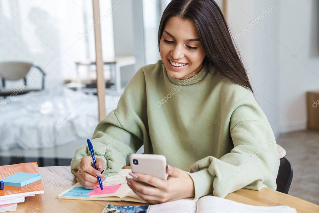 Photo of cheerful beautiful student woman smiling and using cellphone while doing homework in living room