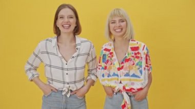 İki mutlu güzel kadın, sarı arka planda kolları ceplerinde eğleniyorlar.