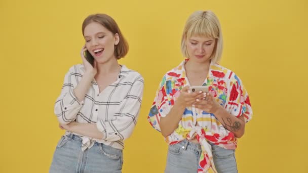 一个快乐的年轻女人站在一个微笑的女人旁边 一边用智能手机在黄色的背景下说话 一边用智能手机和她说话 — 图库视频影像