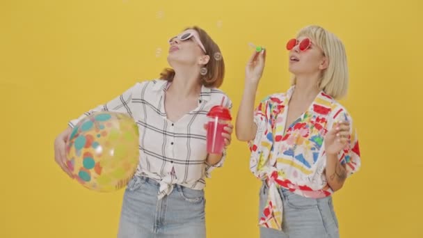 Две Счастливые Красивые Женщины Солнечных Очках Веселятся Мыльными Пузырями Желтом — стоковое видео