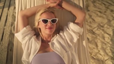 Güneş gözlüğü takan genç ve mutlu bir sarışının manzarası sabahın köründe kumsaldaki hamakta yatıyor.