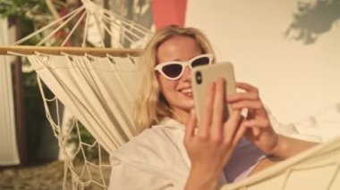 Mutlu, güneş gözlüğü takan genç bir sarışın sabah plajda hamakta yatarken akıllı telefonunu kullanıyor.