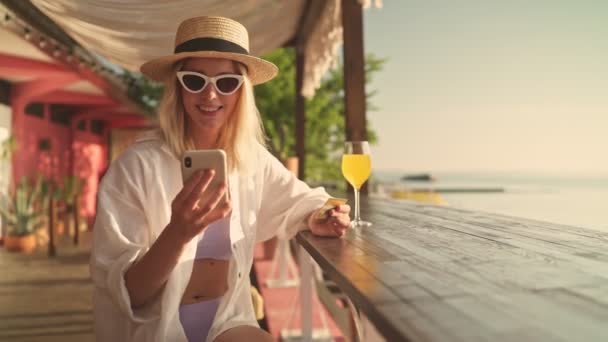 一位戴着太阳镜和帽子的年轻金发女子坐在海滩上的咖啡店里 一边用信用卡和智能手机 一边高兴地看着 — 图库视频影像