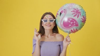 Moda makyajlı ve yüzünde çıkartmalar olan genç bir kız dilek dilerken sarı arka planda duran balonu serbest bırakıyor.
