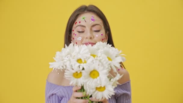 一个满脸都是时尚化妆品和贴纸的快乐而积极的小女孩 手里拿着一束雏菊 孤零零地站在黄色的背景上 — 图库视频影像