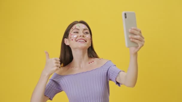 一位满脸都是时尚化妆品和贴纸的情绪激动的年轻姑娘 站在黄色背景下 用智能手机拍下了自己的照片 — 图库视频影像