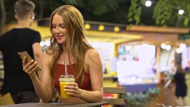 美しい若い女性が夏に市内公園で新鮮なオレンジジュースを飲んでいる間彼女の携帯電話を使用しています — ストック動画