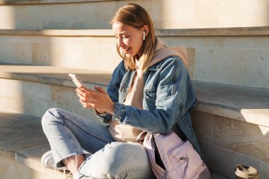 Kapıda otururken cep telefonu kullanan neşeli sarışın kız.