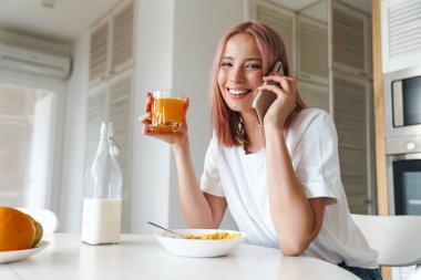 Beyaz mutfakta kahvaltı ederken pembe saçlı, cep telefonuyla konuşan neşeli genç bir kadının fotoğrafı.