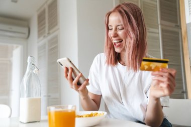 Beyaz mutfakta kahvaltı ederken elinde kredi kartı ve cep telefonu olan genç bir kadının fotoğrafı.