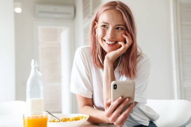 Güzel, neşeli bir kadının beyaz mutfakta kahvaltı ederken cep telefonu ve kulaklık kullanmasının fotoğrafı.