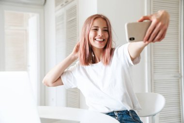 Beyaz mutfakta cep telefonuyla gülümseyen ve selfie çeken beyaz kadın fotoğrafı.
