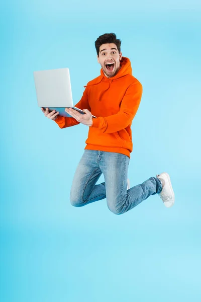 衝撃的な感情的な若い毛の男の写真青の壁の背景の上に隔離ジャンプし ラップトップコンピュータを使用して — ストック写真