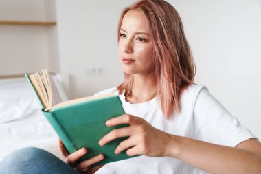 Pembe saçlı, kitap okuyan ve evde dinlenirken düşünen odaklanmış güzel bir kadın resmi.