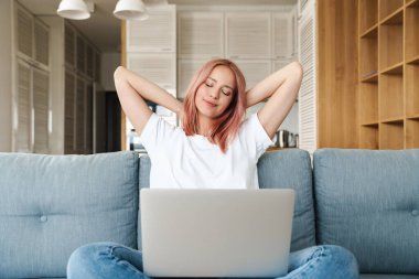 Evdeki kanepede dizüstü bilgisayarla çalışırken pembe saçlı güzel, rahat bir kadının görüntüsü.