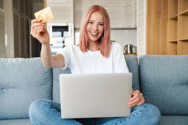 素敵な若い魅力的な女性は リビングルームのソファでリラックスし ラップトップコンピュータを使用して クレジットカードを示しています — ストック写真