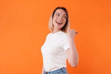Gülümseyen ve işaret eden güzel bir kadının portakal rengi arka planda izole edilmiş görüntüsü.