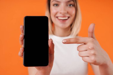 Portakal rengi arka planda izole edilmiş cep telefonunu gösteren ve işaret eden neşeli bir kadın resmi