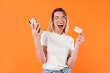 Heyecanlı bir kadının kredi kartını tutarken turuncu arka planda izole edilmiş cep telefonu kullanması.