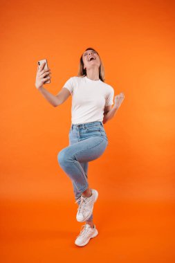 Heyecanlı bir kadının, turuncu arka planda izole edilmiş cep telefonu kullanarak birinci sınıf jest yapması.