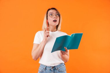 Düşünceli genç bir kadının portresi günlük kıyafetlerle turuncu arka planda izole bir şekilde duruyor, bir günlüğe not alıyor, başka tarafa bakıyor.