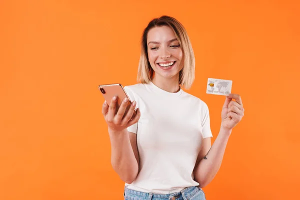 オレンジの背景に隔離された携帯電話を使用しながらクレジットカードを保持して幸せな素敵な女性のイメージ — ストック写真