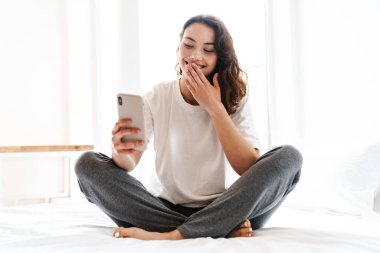 Evde yatakta otururken gülümseyen ve cep telefonu kullanan neşeli, çekici bir kadının fotoğrafı.