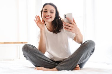 Neşeli, çekici bir kadının el salladığı ve evde otururken cep telefonu kullandığı bir fotoğraf.