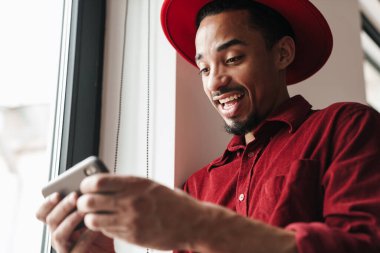 Pencerenin yanında cep telefonu kullanan yakışıklı, mutlu ve genç bir adamın görüntüsü