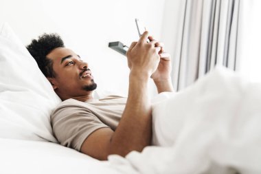 Beyaz yatakta cep telefonu kullanan, bıyıklı, gülümseyen Afrikalı Amerikalı adamın fotoğrafı.